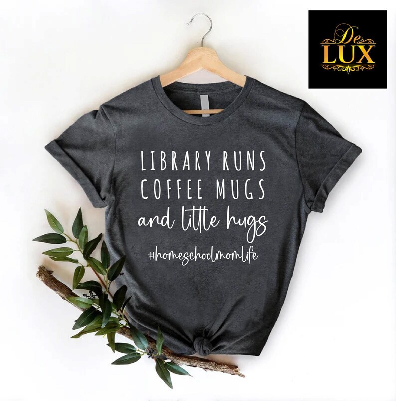 May include: Dark gray t-shirt with the text "LIBRARY RUNS COFFEE MUGS and little hugs" in white. The hashtag "#homeschoolmomlife" is below the text. The shirt is hanging on a wooden hanger.