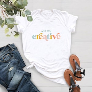 May include: White t-shirt with the text "let's get creative" in colorful, retro-style lettering. The shirt is paired with blue denim jeans and brown sandals with black straps. The items are arranged on a light-colored surface.