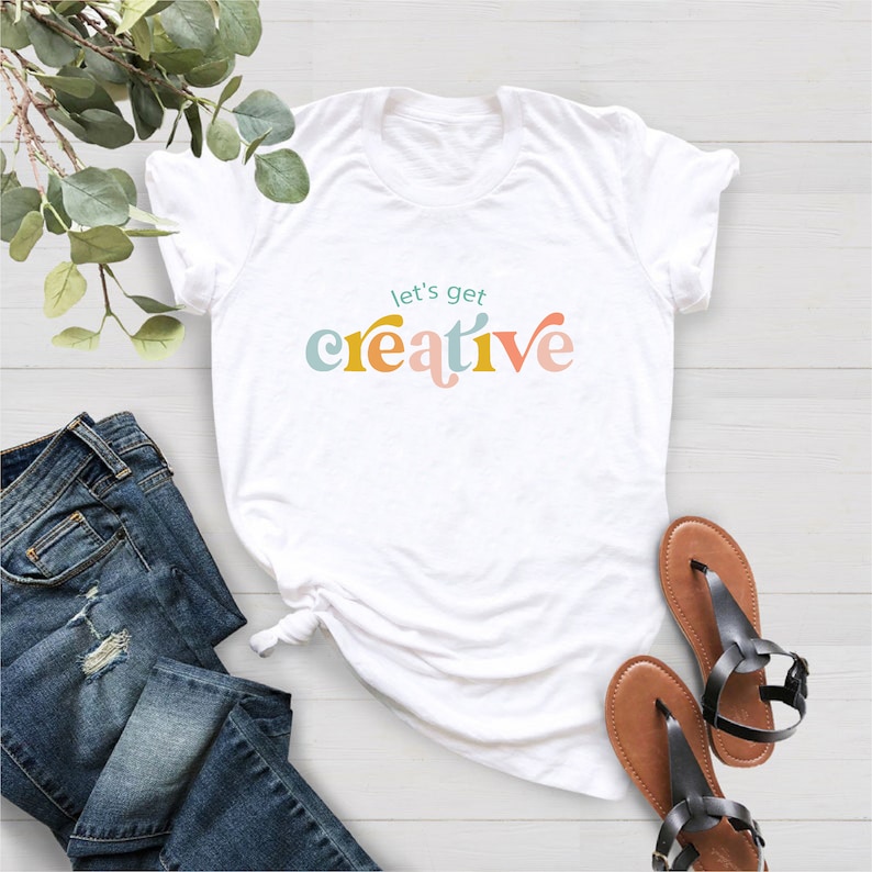 May include: White t-shirt with the text "let's get creative" in colorful, retro-style lettering. The shirt is paired with blue denim jeans and brown sandals with black straps. The items are arranged on a light-colored surface.