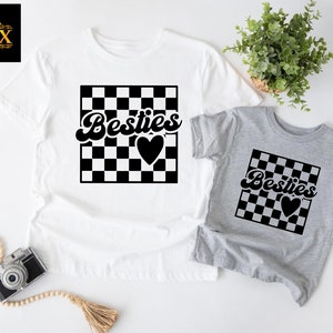 K&ouml;nnte beinhalten: Zwei T-Shirts, eines wei&szlig; und eines grau, mit passendem Design. Jedes Shirt hat ein schwarz-wei&szlig; kariertes Quadrat mit dem Wort "Besties" in Schreibschrift und einem schwarzen Herzen. Das wei&szlig;e Shirt ist gr&ouml;&szlig;er als das graue Shirt.