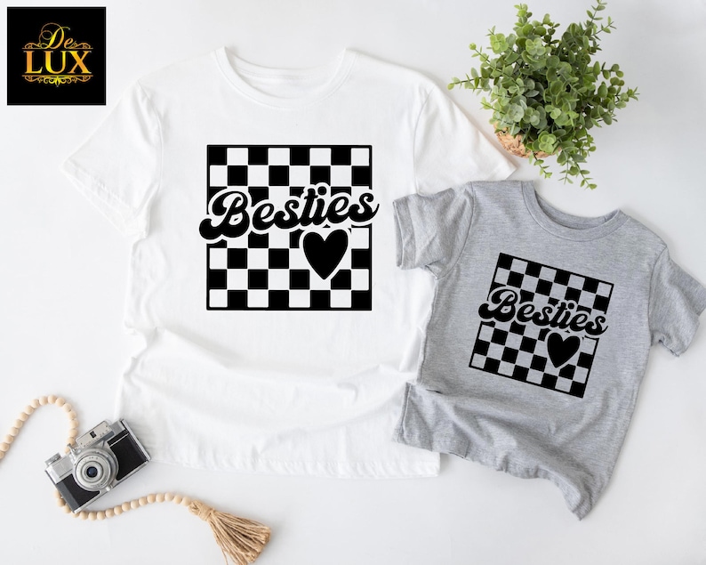 K&ouml;nnte beinhalten: Zwei T-Shirts, eines wei&szlig; und eines grau, mit passendem Design. Jedes Shirt hat ein schwarz-wei&szlig; kariertes Quadrat mit dem Wort "Besties" in Schreibschrift und einem schwarzen Herzen. Das wei&szlig;e Shirt ist gr&ouml;&szlig;er als das graue Shirt.