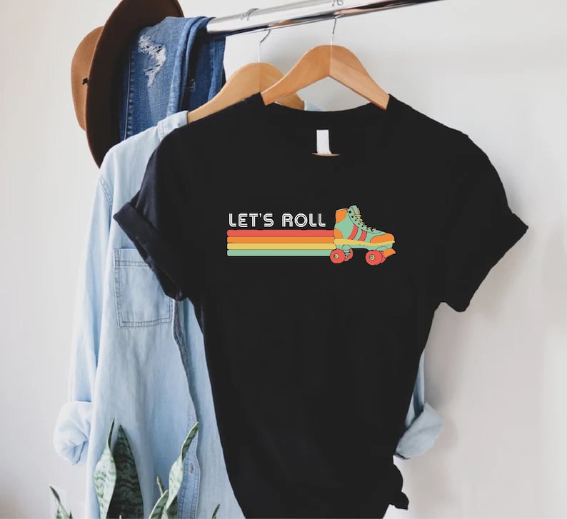 May include: Black t-shirt with the text "LET'S ROLL" above a retro roller skate graphic. The graphic features a teal and orange roller skate with red wheels, and a horizontal stripe design in orange, yellow, and green.