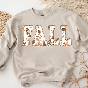 May include: A beige sweatshirt with the word "FALL" in large, block letters. The letters are filled with a pattern of fall leaves, acorns, and other autumn-themed designs in shades of brown and white.