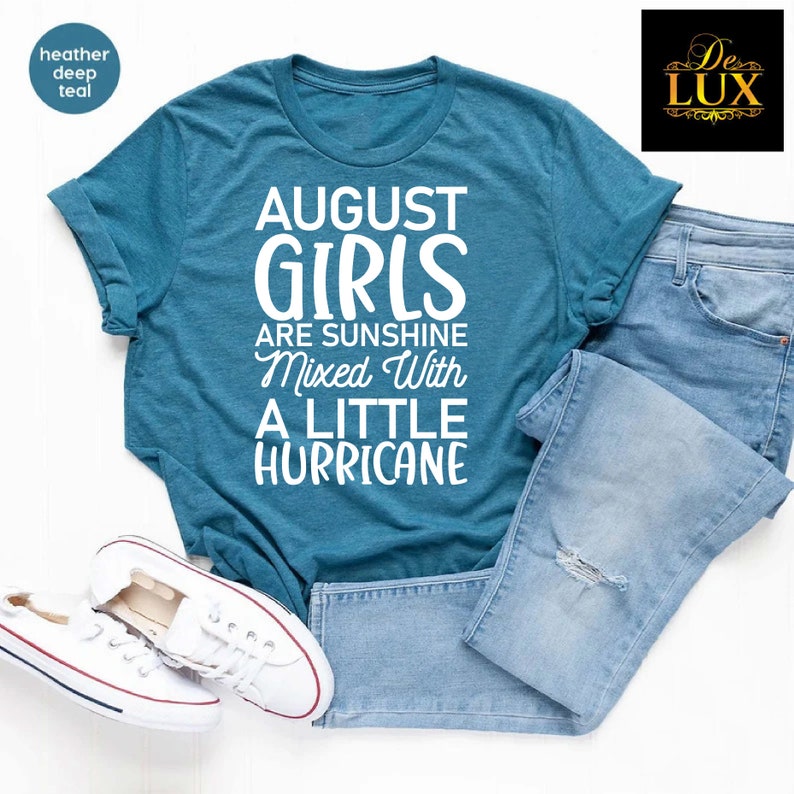 May include: A teal t-shirt with the text "AUGUST GIRLS ARE SUNSHINE MIXED WITH A LITTLE HURRICANE" in white. The shirt is paired with light blue jeans and white sneakers.
