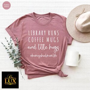 May include: Mauve-colored t-shirt with the text "LIBRARY RUNS COFFEE MUGS and little hugs #homeschoolmomlife". A white coffee mug and a beige hat are next to the shirt. The shirt is folded and tied at the bottom.