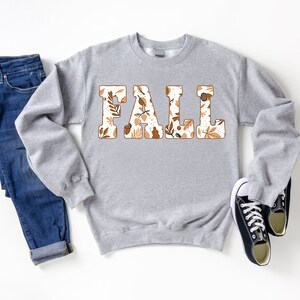 May include: A gray sweatshirt with the word "FALL" in large, block letters. The letters are filled with a pattern of fall leaves and acorns in shades of brown and cream. Dark blue jeans and black canvas sneakers complete the fall-themed ensemble.