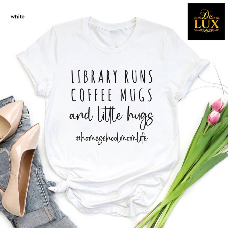May include: White t-shirt with the text "LIBRARY RUNS COFFEE MUGS and little hugs #homeschoolmomlife" in black. The shirt is styled with a pair of beige heels and a pair of blue jeans. A logo is in the upper right corner.