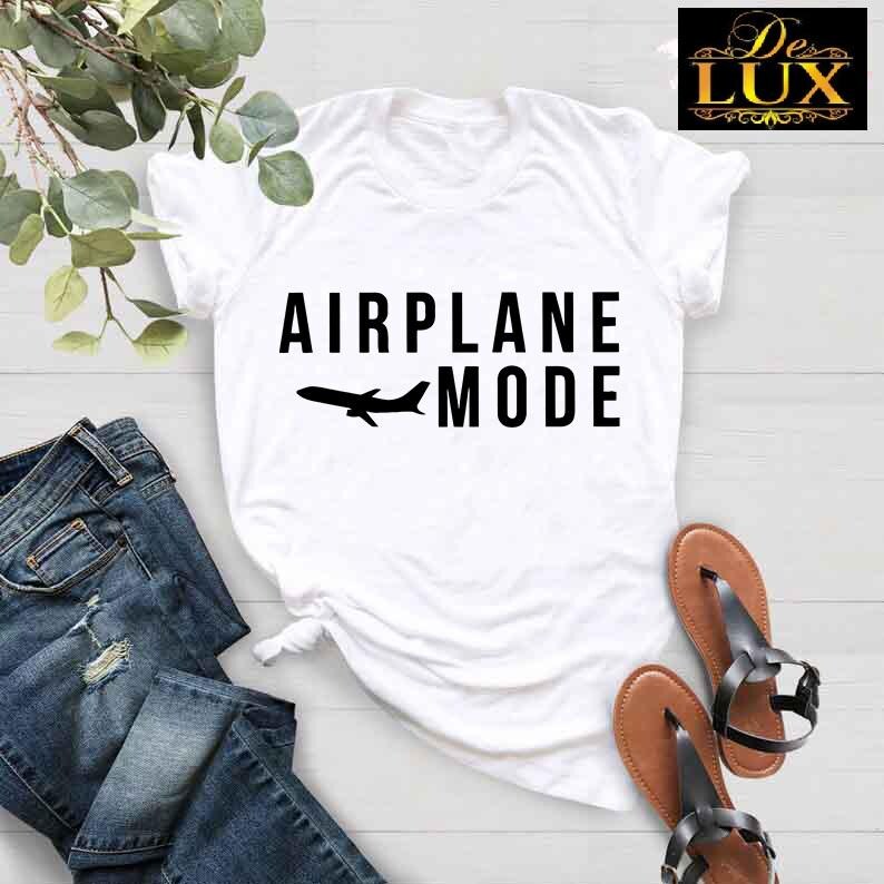 Puede incluir: Camiseta blanca con la frase "AIRPLANE MODE" en negro y un avi&oacute;n negro. La imagen muestra la camiseta con vaqueros azules y sandalias marrones. Un logotipo es visible en la esquina superior derecha.