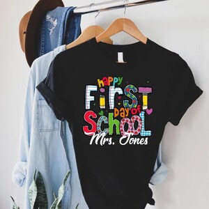 May include: Black t-shirt with the text "happy First day of School Mrs. Jones" in colorful letters. The design includes pencils, hearts, and a notebook-style font. The shirt is hanging on a wooden hanger.
