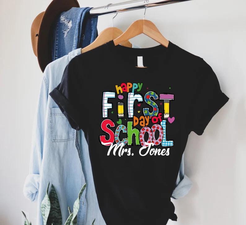 May include: Black t-shirt with the text "happy First day of School Mrs. Jones" in colorful letters. The design includes pencils, hearts, and a notebook-style font. The shirt is hanging on a wooden hanger.