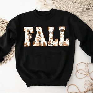 May include: Black sweatshirt with the word "FALL" in large, block letters. The letters are filled with a pattern of fall leaves and acorns in shades of brown and cream. The sweatshirt has long sleeves.