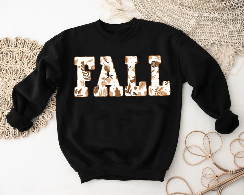 May include: Black sweatshirt with the word "FALL" in large, block letters. The letters are filled with a pattern of fall leaves and acorns in shades of brown and cream. The sweatshirt has long sleeves.