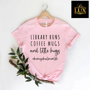May include: A light pink t-shirt with the text "LIBRARY RUNS COFFEE MUGS and little hugs" in black. The hashtag "#homeschoolmomlife" is below the text. The shirt is hanging on a wooden hanger.