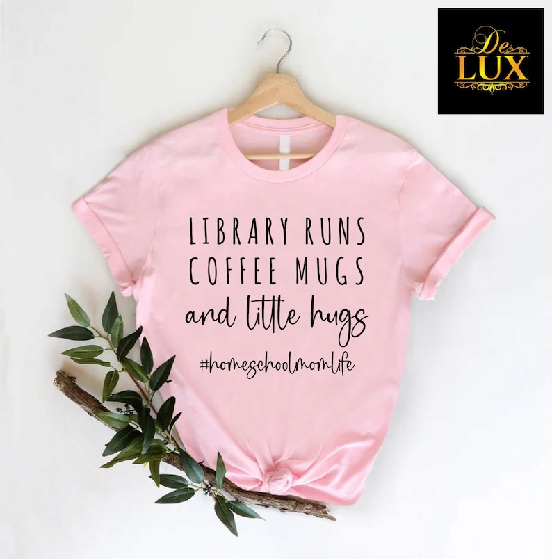 May include: A light pink t-shirt with the text "LIBRARY RUNS COFFEE MUGS and little hugs" in black. The hashtag "#homeschoolmomlife" is below the text. The shirt is hanging on a wooden hanger.
