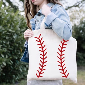 May include: A cream-colored tote bag featuring a baseball design. Two red, curved lines, mimicking the stitching of a baseball, are printed on the front. The bag is held by a person wearing a denim jacket.