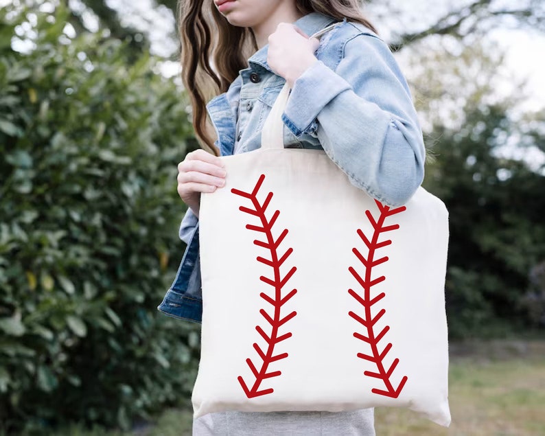 May include: A cream-colored tote bag featuring a baseball design. Two red, curved lines, mimicking the stitching of a baseball, are printed on the front. The bag is held by a person wearing a denim jacket.