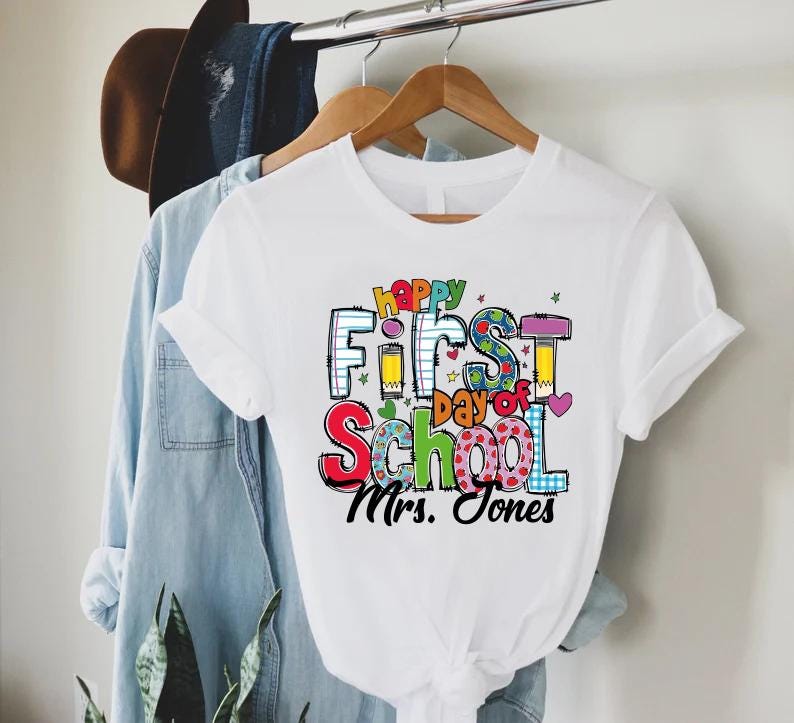 May include: White t-shirt with the text "Happy First Day of School Mrs. Jones" in colorful block letters. The shirt is hanging on a wooden hanger with a denim shirt and a brown hat in the background. The design includes hearts, stars, and pencils.