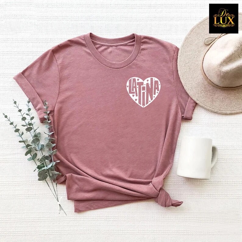 May include: A dusty rose t-shirt with a white heart graphic that says "LATINA". The t-shirt has rolled-up sleeves and a knotted hem. A white mug and a beige hat are also in the image.