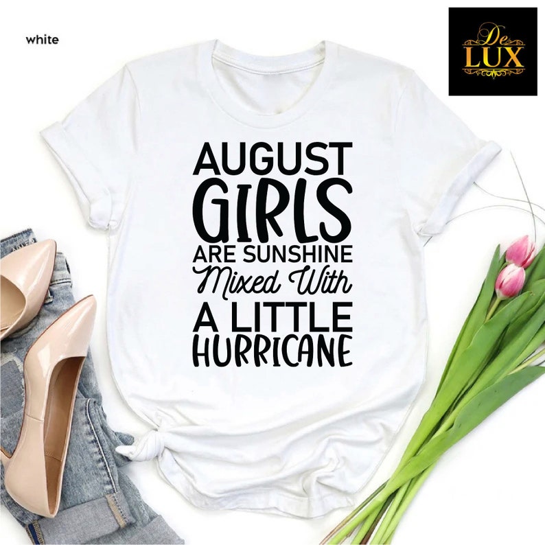 May include: White t-shirt with the text "AUGUST GIRLS ARE SUNSHINE Mixed With A LITTLE HURRICANE" in black. The shirt is styled with a pair of beige heels, blue jeans, and a bouquet of pink tulips.