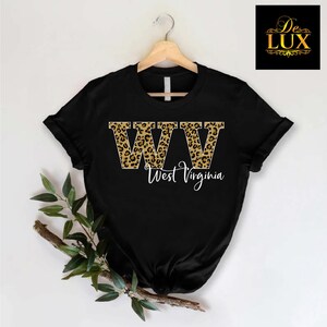 May include: Black t-shirt with the letters "WV" in a leopard print design, with "West Virginia" written below in a cursive font. The shirt is hanging on a wooden hanger.