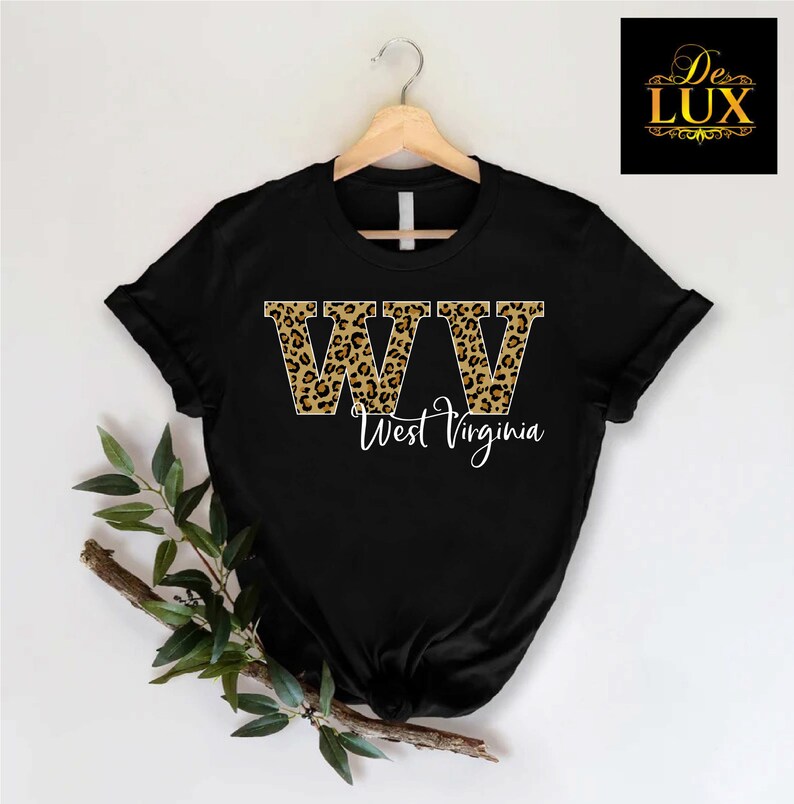 May include: Black t-shirt with the letters "WV" in a leopard print design, with "West Virginia" written below in a cursive font. The shirt is hanging on a wooden hanger.