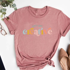 May include: A dusty rose-colored t-shirt with the phrase "let's get creative" printed across the front in a colorful, retro font. The shirt has short sleeves and a crew neck. The bottom of the shirt is tied in a knot.