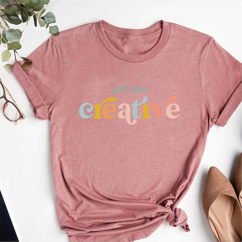 May include: A dusty rose-colored t-shirt with the phrase "let's get creative" printed across the front in a colorful, retro font. The shirt has short sleeves and a crew neck. The bottom of the shirt is tied in a knot.