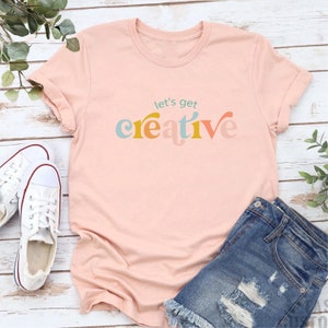 May include: A light pink t-shirt with the words "let's get creative" in colorful text. The shirt is paired with denim shorts and white canvas shoes. The shirt has rolled-up sleeves.