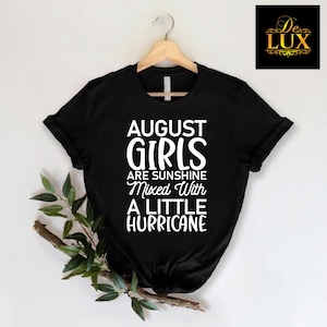May include: Black t-shirt with the text "AUGUST GIRLS are sunshine mixed with a little hurricane" in white. The shirt is hanging on a wooden hanger. A sprig of greenery is on the left side of the shirt.