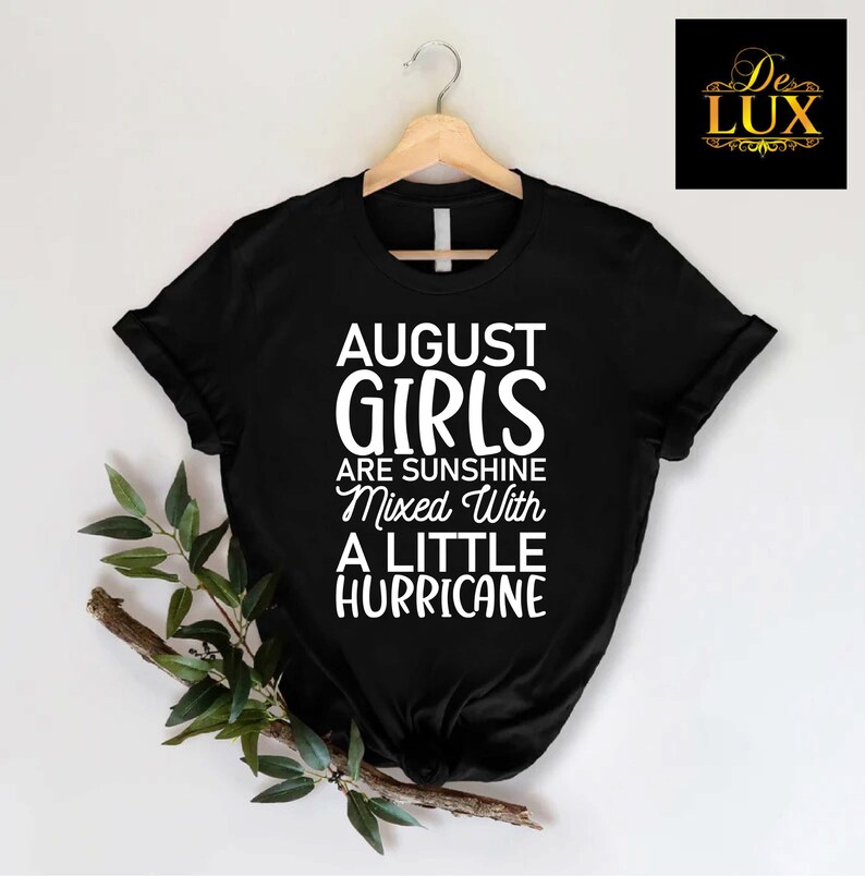 May include: Black t-shirt with the text "AUGUST GIRLS are sunshine mixed with a little hurricane" in white. The shirt is hanging on a wooden hanger. A sprig of greenery is on the left side of the shirt.