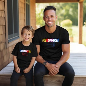 May include: Two people wearing matching black t-shirts with the text "SHAKE" and "& BAKE" in a colorful design. The shirts feature a fist graphic and a racing stripe design. The people are sitting on a wooden porch.