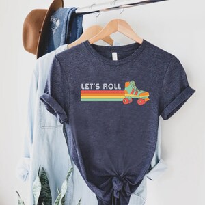 May include: Navy blue t-shirt with the text "LET'S ROLL" above a retro design of a roller skate and a horizontal stripe in red, orange, yellow, and green. The shirt is hanging on a wooden hanger.