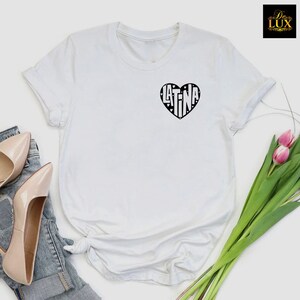 May include: White t-shirt featuring a black heart-shaped design with the word "LATINA" inside. The shirt has short sleeves and a crew neck. The image also shows a pair of beige high heels, denim jeans, and a bouquet of pink tulips.