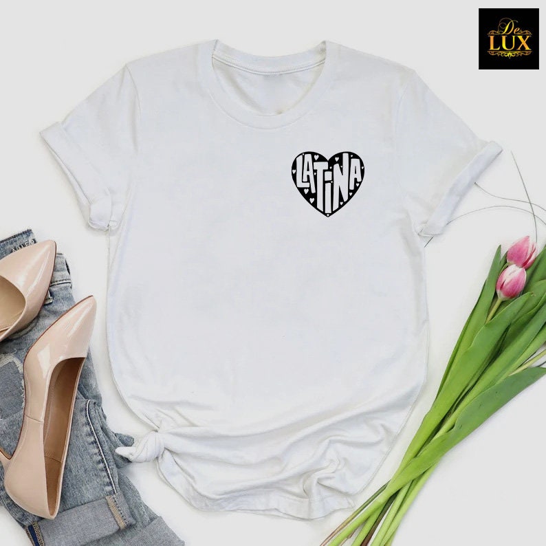May include: White t-shirt featuring a black heart-shaped design with the word "LATINA" inside. The shirt has short sleeves and a crew neck. The image also shows a pair of beige high heels, denim jeans, and a bouquet of pink tulips.