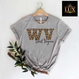 May include: A heather gray t-shirt featuring the letters "WV" in a leopard print design, with "West Virginia" written below in a script font. The shirt is displayed on a wooden hanger.