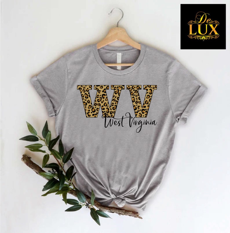 May include: A heather gray t-shirt featuring the letters "WV" in a leopard print design, with "West Virginia" written below in a script font. The shirt is displayed on a wooden hanger.