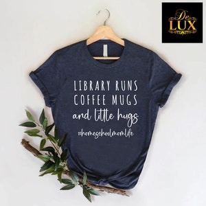 May include: Navy blue t-shirt with the text "LIBRARY RUNS COFFEE MUGS and little hugs" in white script. The hashtag "#homeschoolmomlife" is below the text. The shirt is hanging on a wooden hanger.