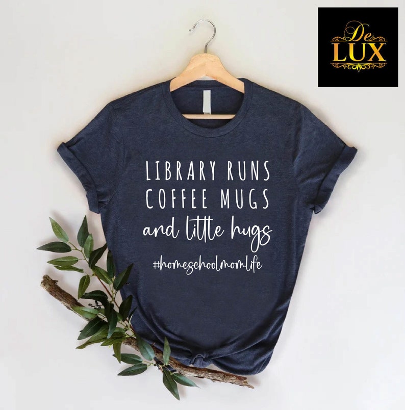 May include: Navy blue t-shirt with the text "LIBRARY RUNS COFFEE MUGS and little hugs" in white script. The hashtag "#homeschoolmomlife" is below the text. The shirt is hanging on a wooden hanger.