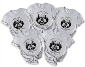 May include: Five light gray short-sleeve t-shirts featuring a black graphic of a tent, campfire, pine trees, and a crescent moon. The text reads Groveland, CA, Wilson Family, and 2026. The shirts are arranged in a cluster with two tied at the hem.