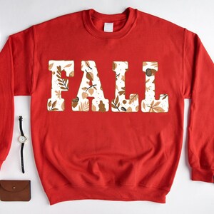 May include: A red sweatshirt with the word "FALL" in large, block letters. The letters are filled with a pattern of fall leaves, acorns, and other autumn-themed designs. The sweatshirt is displayed on a white surface.