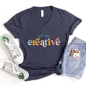 May include: Navy blue V-neck t-shirt with the text "let's get creative" in colorful, playful lettering. The shirt is paired with denim shorts, gray sneakers, and a floral phone case. A casual, artistic theme.