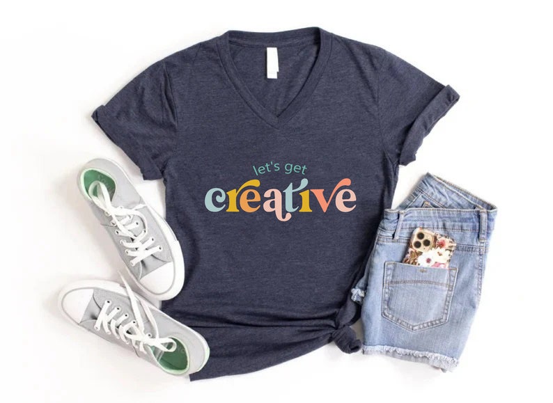 May include: Navy blue V-neck t-shirt with the text "let's get creative" in colorful, playful lettering. The shirt is paired with denim shorts, gray sneakers, and a floral phone case. A casual, artistic theme.