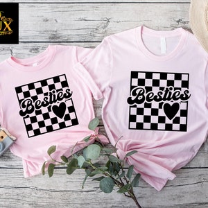 K&ouml;nnte beinhalten: Zwei hellrosa T-Shirts. Jedes zeigt das Wort "Besties" in schwarzer Schreibschrift, &uuml;ber einem schwarz-wei&szlig; karierten Quadrat und einem schwarzen Herz. Die Shirts sind ein passendes Set, eines kleiner als das andere.