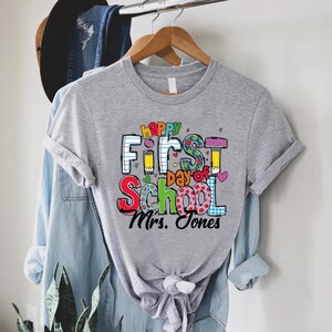 May include: A gray t-shirt with the words "Happy First Day of School" in colorful letters. The shirt also includes the name "Mrs. Jones" in black script. The shirt is hanging on a wooden hanger.