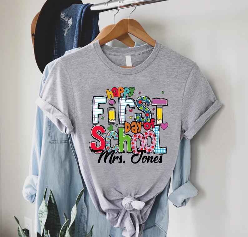 May include: A gray t-shirt with the words "Happy First Day of School" in colorful letters. The shirt also includes the name "Mrs. Jones" in black script. The shirt is hanging on a wooden hanger.