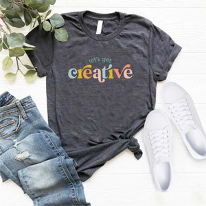 May include: Dark gray t-shirt with the phrase "let's get creative" in colorful, retro-style lettering. The shirt is paired with blue jeans and white sneakers, suggesting a casual, artistic theme.