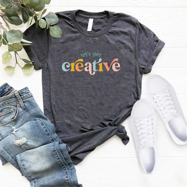 May include: Dark gray t-shirt with the phrase "let's get creative" in colorful, retro-style lettering. The shirt is paired with blue jeans and white sneakers, suggesting a casual, artistic theme.