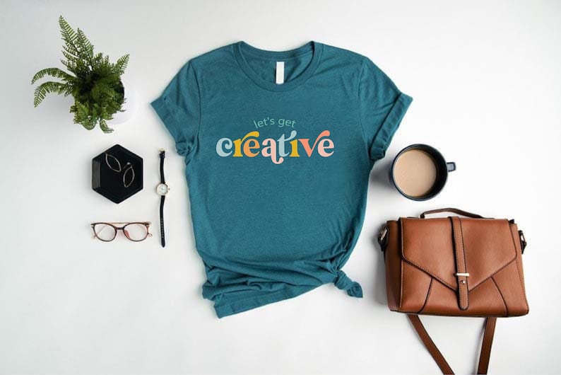 May include: Teal t-shirt with the phrase "let's get creative" in colorful, stylized lettering. The shirt is styled with a watch, glasses, a black tray with earrings, a plant, a coffee cup, and a brown satchel.