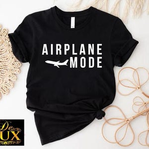 Puede incluir: Camiseta negra con las palabras "AIRPLANE MODE" en blanco y un avi&oacute;n blanco. La camiseta est&aacute; anudada en la parte inferior. El logotipo de De Lux est&aacute; en la esquina inferior izquierda.