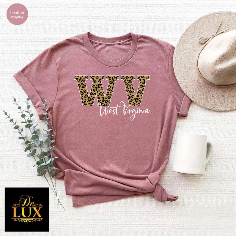 May include: Mauve-colored t-shirt featuring the letters "WV" in a leopard print design, with "West Virginia" written below in a cursive font. The shirt is styled with a hat and a white mug.
